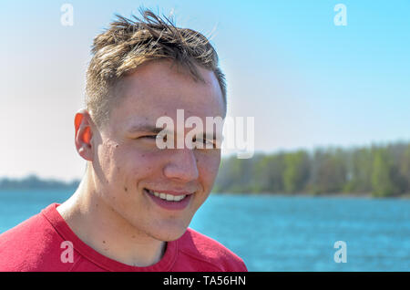 Demi-tour buste portrait d'un jeune gars en t-shirt rouge, debout à l'extérieur, sur le lac ou la rivière. Looking at camera and smiling Banque D'Images