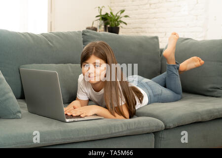 Beau charmant mignon lycéenne jouer et surfer sur l'Internet sur ordinateur portable en souriant et en étant couché sur le canapé à la maison. La technologie numérique, interne Banque D'Images