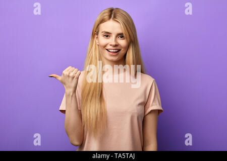 Jeune fille charmante optimiste avec sourire à pleines dents à l'aide du pouce de côté points, spectacles de l'espace pour la publicité. close up portrait.hurry up Banque D'Images