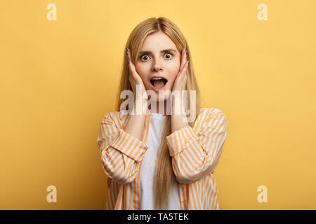 Juste magnifique émotionnel-haired woman screaming avec choc, en se tenant par la main sur ses joues. close up photo. fond jaune isolé. studio shot.emot Banque D'Images