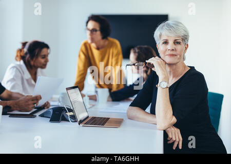 Femme d'affaires à maturité dans la salle de réunion avec des collègues discutant en arrière-plan. Confident female professionnel à bord prix réunion. Banque D'Images