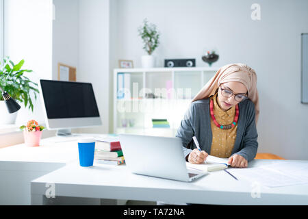 Jeune enseignante musulmane assis près de l'ordinateur portable et la rédaction de notes Banque D'Images