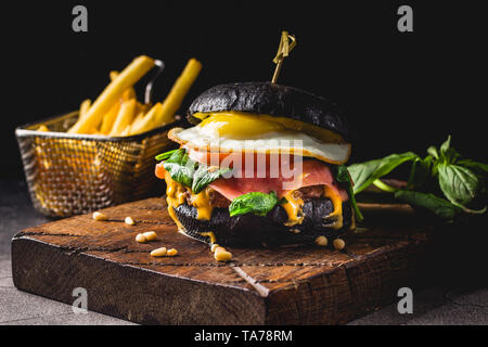 Burger de boeuf avec black bun, fromage, oeufs brouillés, jambon, tomates, roquette et chard et frites sur une planche à découper en bois. Street food, restauration rapide foo Banque D'Images