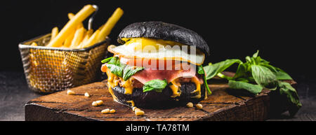 Burger de boeuf avec black bun, fromage, oeufs brouillés, jambon, tomates, roquette et chard et frites sur une planche à découper en bois. Street food, restauration rapide foo Banque D'Images