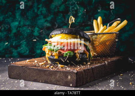 Burger de boeuf avec black bun, fromage, oeufs brouillés, jambon, tomates, roquette et chard et frites sur une planche à découper en bois. Street food, restauration rapide foo Banque D'Images