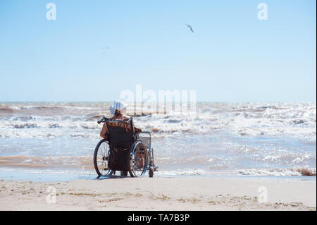 Vue arrière d'une femme handicapée assise sur un fauteuil roulant sur la plage par temps ensoleillé. Elle regarde l'océan Banque D'Images