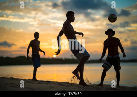 Fernando Machado, BRÉSIL - Février 2018 : les jeunes hommes jouent un jeu informel de beach-football-keepy uppy en silhouette sur le rivage d'une plage au coucher du soleil. Banque D'Images