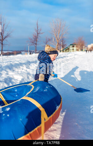 Un petit garçon de 3 ans, en hiver sur rue est en faisant glisser les tubes, pour l'équitation d'une colline, dans les mains d'un terrain de paddle, jouer, s'amuser Banque D'Images