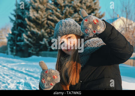 Belle jeune fille de race blanche avec les cheveux dans un bonnet, écharpe en tricot et mitaines en plein air dans le village sur un après-midi après-midi d'hiver montre une bonne humeur Banque D'Images