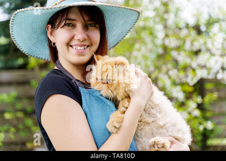 Photo de happy woman in hat avec chat dans les mains sur fond flou dans jardin Banque D'Images