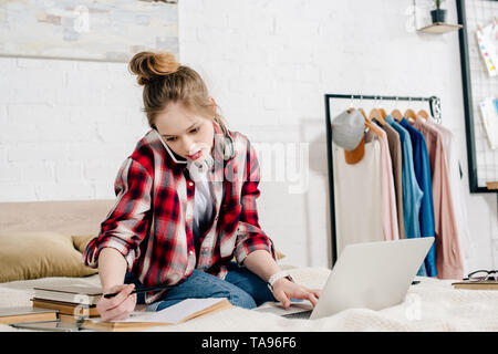 Adolescent inquiet assis sur le lit avec des livres et d'ordinateurs portables et de parler sur le smartphone. Banque D'Images