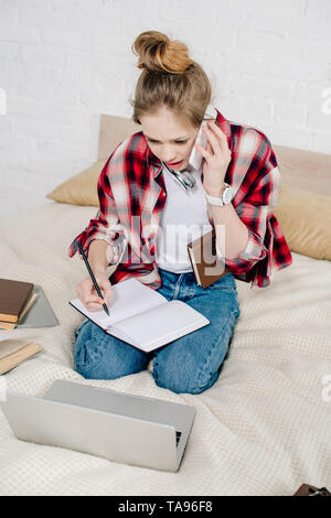 Adolescent inquiet assis sur le lit avec des livres et d'ordinateurs portables et de parler sur le smartphone. Banque D'Images