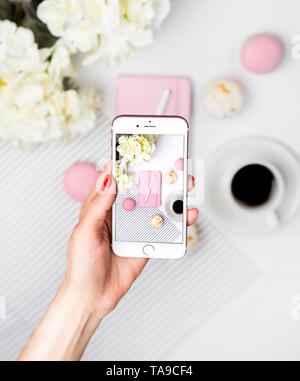 Femme prendre une photo d'espace de travail sur un smartphone. Ordinateur portable rose, un stylo, de macarons, de café et de fleurs sur fond blanc. Bureau des femmes, bureau, Banque D'Images