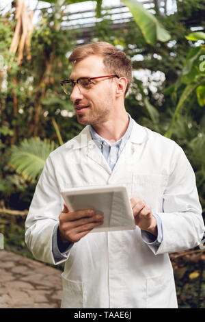 Smiling handsome scientifique en blouse blanche et verres using digital tablet in orangerie Banque D'Images