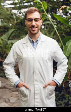 Smiling handsome scientifique en blouse blanche et verres debout avec les mains dans les poches à l'orangerie Banque D'Images