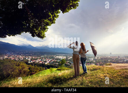 Deux amis avec parapluie à la recherche de la belle vue de panorama city dans les montagnes au coucher du soleil spectaculaire des pluies Banque D'Images