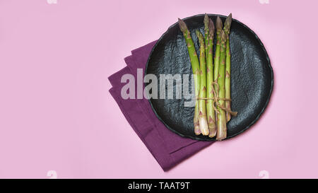 Les asperges vertes fraîches sur plat de céramique noire, violet , serviette et fond rose. Banque D'Images