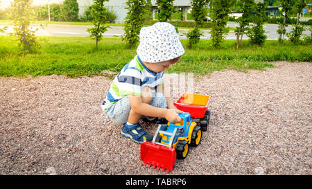 Portrait of cute 3 ans Bébé garçon assis sur l'aire de jeux au parc et jouer avec des jouets en plastique chariot. Enfant s'amuser et jouer Banque D'Images