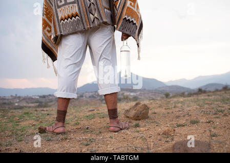 L'homme de derrière debout sur un sol sec portant un pantalon blanc / pantalon, tapis anciens, zapotèque et sandales une bouteille de mezcal. L'État de Oaxaca, Mexique Banque D'Images