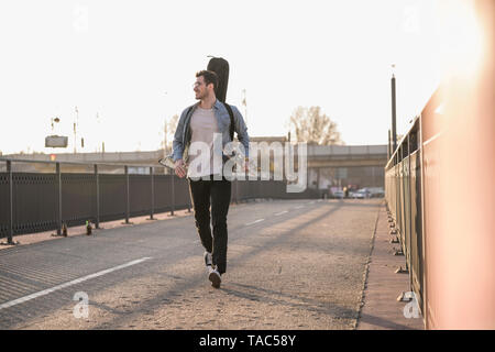 Smiling Young man with guitar case et skateboard marche sur un pont dans la ville Banque D'Images