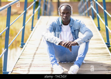 Portrait d'un homme portant des vêtements en jean décontracté assis sur la passerelle à l'écoute de la musique avec des écouteurs Banque D'Images