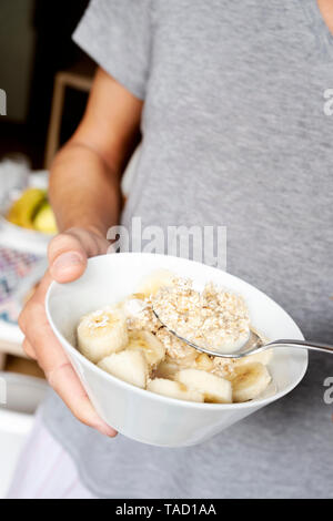 Libre d'un jeune homme portant un T-shirt gris occasionnels ayant un peu de porridge à la banane d'un bol en céramique blanche, debout à côté d'une table définie pour brea Banque D'Images