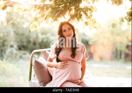 Pregnant woman wearing dress posing rose élégant en plein air avec le coucher du soleil sur l'arrière-plan. En regardant la caméra. La maternité. La maternité. Banque D'Images