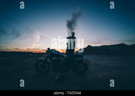 Homme barbu avec des dreadlocks assis sur moto au coucher du soleil de fumer cigarette électronique Banque D'Images