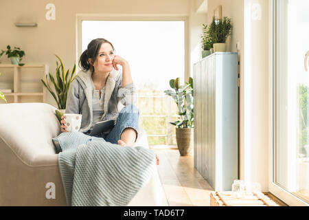 Femme souriante avec une tasse et tablet assis sur le canapé à la maison Banque D'Images