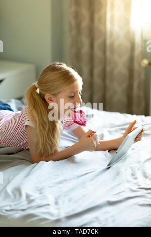 Blonde girl lying on bed licking lollipop lors de l'utilisation de digital tablet Banque D'Images