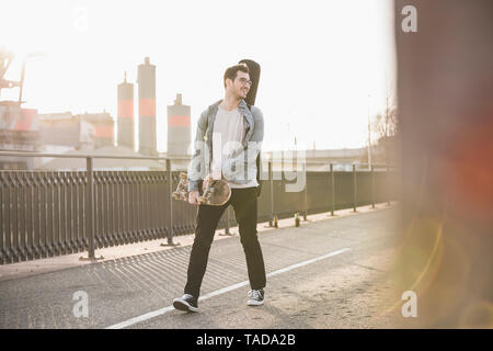 Smiling Young man with guitar case et skateboard marche sur un pont dans la ville Banque D'Images