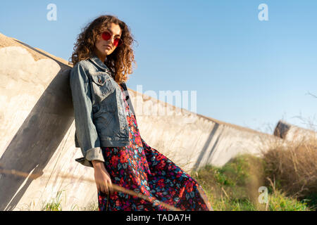 Portrait de jeune femme aux cheveux bruns bouclés portant des lunettes rouge leaning against wall Banque D'Images