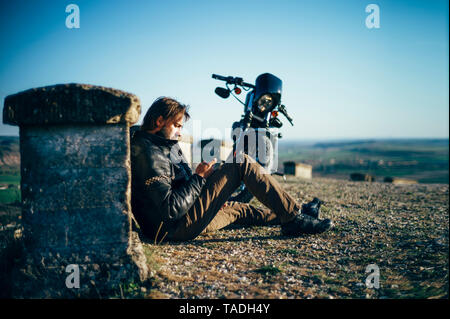 L'homme avec la motocyclette dont une pause assis sur le sol à l'aide de téléphone cellulaire Banque D'Images