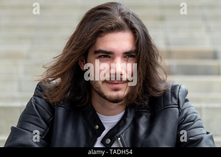 Portrait d'un bel homme barbu avec de longs cheveux bruns, habillé dans les vêtements à la mode, assis sur les marches et à la recherche sur le côté, avec pour zone de contenu publicitaire. Hipster beau mec posant sur l'exemplaire de l'espace. Banque D'Images