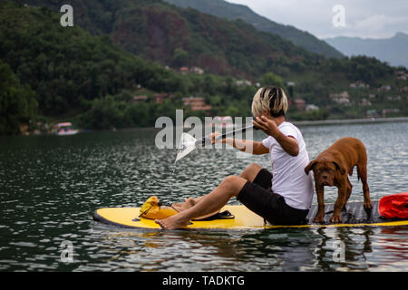 Jeune homme de l'aviron sur paddleboard accompagné de chien dans le lac de Pokhara au Népal. Phewa Tal Barahi Mandir Lac Phewa Banque D'Images