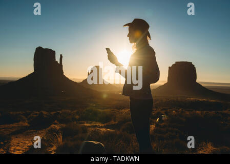 USA, Utah, Monument Valley, silhouette de femme avec chapeau de cow-boy looking at mobile phone at sunrise Banque D'Images