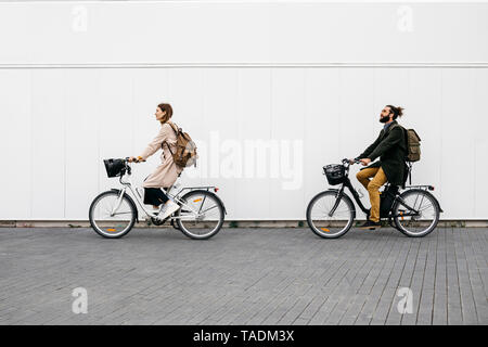 Couple riding e-bikes le long d'un mur blanc dans la ville Banque D'Images