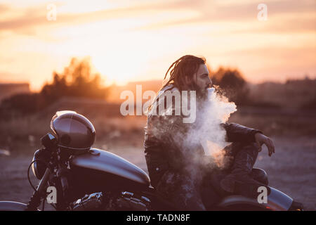 Homme barbu avec des dreadlocks assis sur moto au coucher du soleil de fumer cigarette électronique Banque D'Images