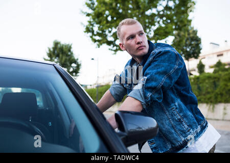 Jeune homme en veste en jean appuyé contre voiture Banque D'Images