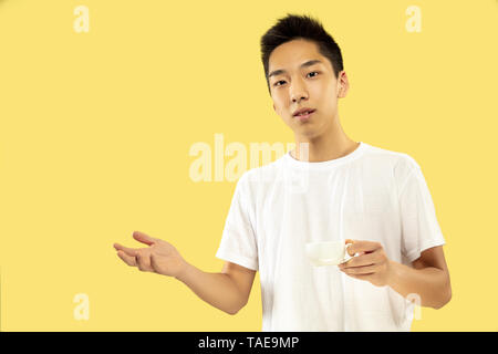 Jeune homme coréen demi-longueur du portrait sur fond jaune studio. Modèle masculin en chemise blanche. Boire du café et invitant. Concept de l'émotion humaine Banque D'Images