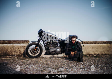 L'homme avec la motocyclette dont une pause assis sur le sol à l'aide de téléphone cellulaire Banque D'Images