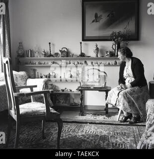 Années 1960, historique, vieille dame assise dans une chambre à l'avant à côté de sa perruche dans une cage, avec des étagères de la knic-knacs et ornements sur le mur derrière, England, UK. Banque D'Images