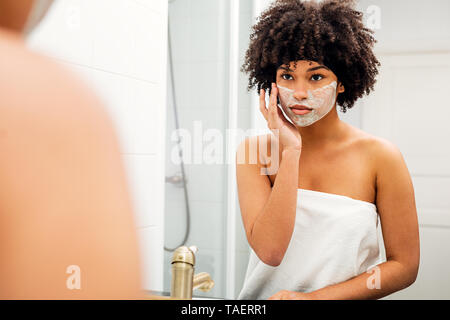 Woman applying facial mask en face du miroir de salle de bains Banque D'Images