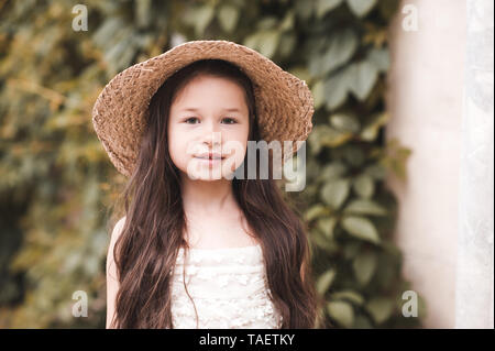 Sourire petite fille 4-5 ans portant chapeau de paille et robe blanche sur fond nature. En regardant la caméra. Vacances d'été. Banque D'Images