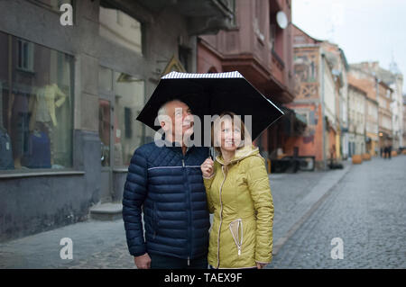 L'extérieur portrait d'un homme âgé et sa jeune femme aux cheveux blonds enlacés debout sous leur parapluie sur rue pavée. Couple avec l'âge Banque D'Images