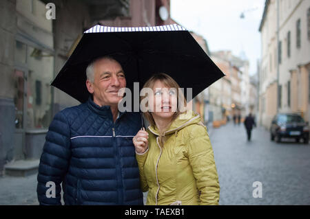 Portrait d'un homme âgé et sa jeune femme aux cheveux blonds enlacés et permanent, sous leur parapluie sur rue pavée. Couple avec Banque D'Images