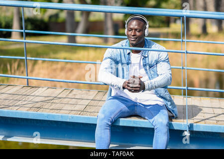 Portrait d'un homme portant des vêtements en jean décontracté assis sur la passerelle d'écouter la musique avec des écouteurs sans fil Banque D'Images