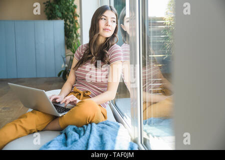 Jeune femme assise à la fenêtre à la maison à l'aide d'ordinateur portable Banque D'Images