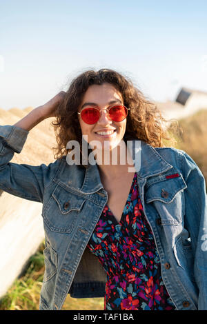 Portrait de jeune femme avec les cheveux bruns bouclés portant des lunettes rouges appuyée sur un mur Banque D'Images