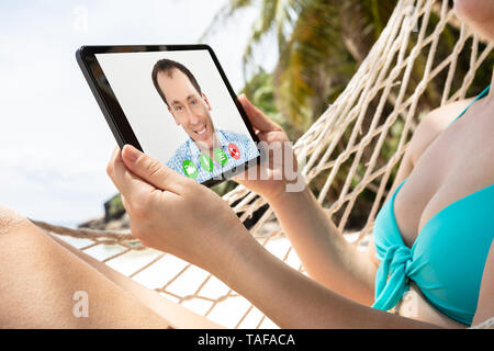 Young Woman Lying On Hammock l'appel vidéo sur tablette numérique avec son petit ami At Beach Banque D'Images